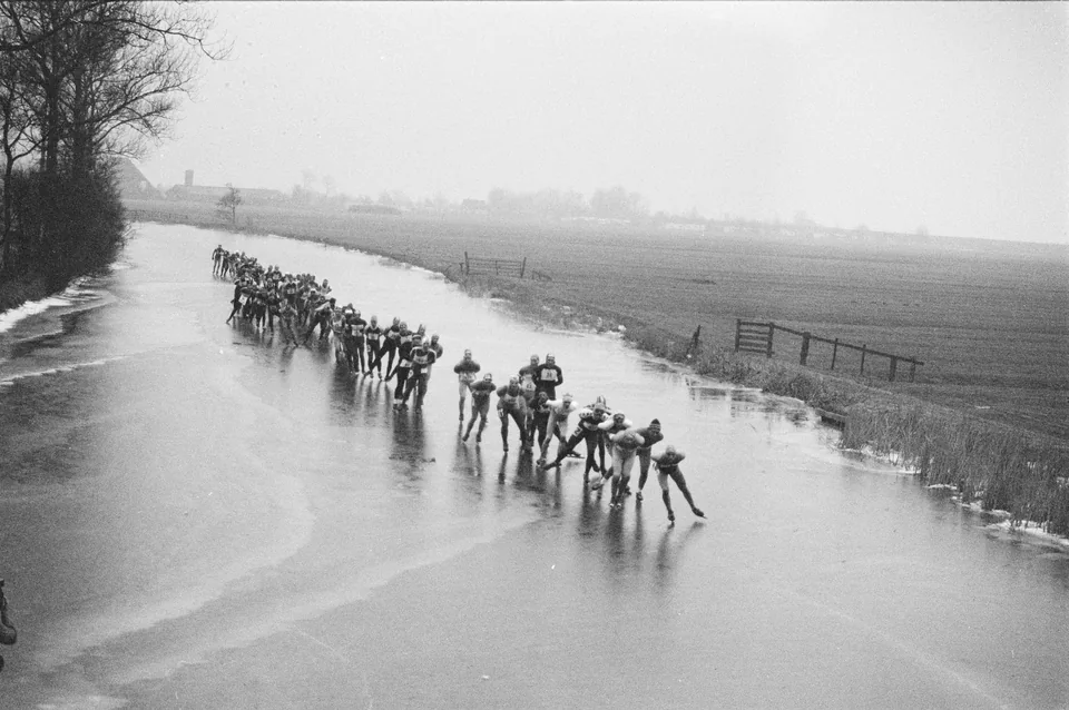 Een lange kopgroep schaatst achter elkaar over een vaart tussen weilanden.
